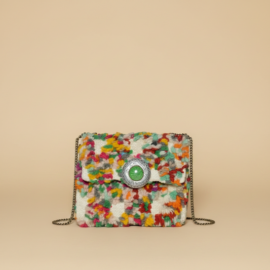 A small flap shoulder bag with a multicolor abstract patterned tweed body, featuring a central silver-tone circular clasp with a green stone and a thin silver chain strap, set against a solid beige background.