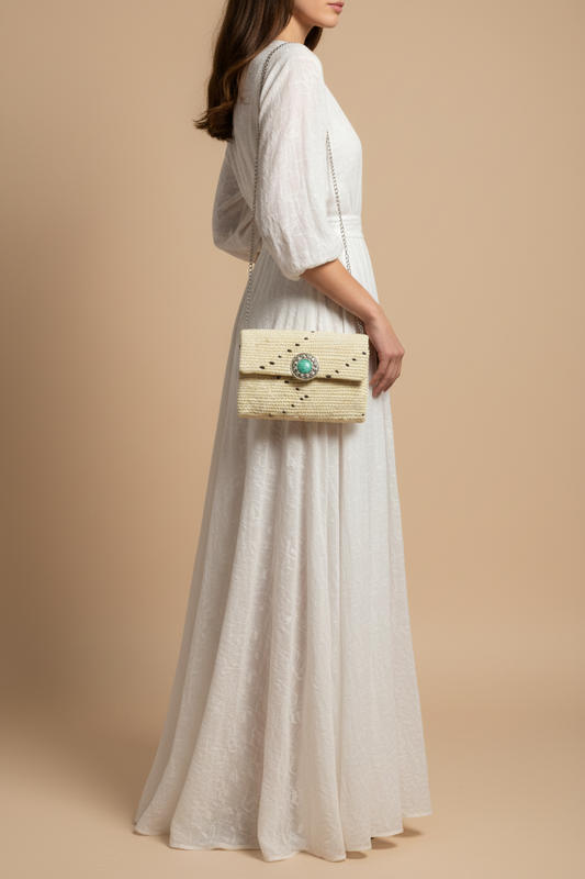 A woman wearing a long white eyelet maxi dress and holding a cream croc-embossed clutch bag with a turquoise stone clasp, posed against a solid beige studio background.