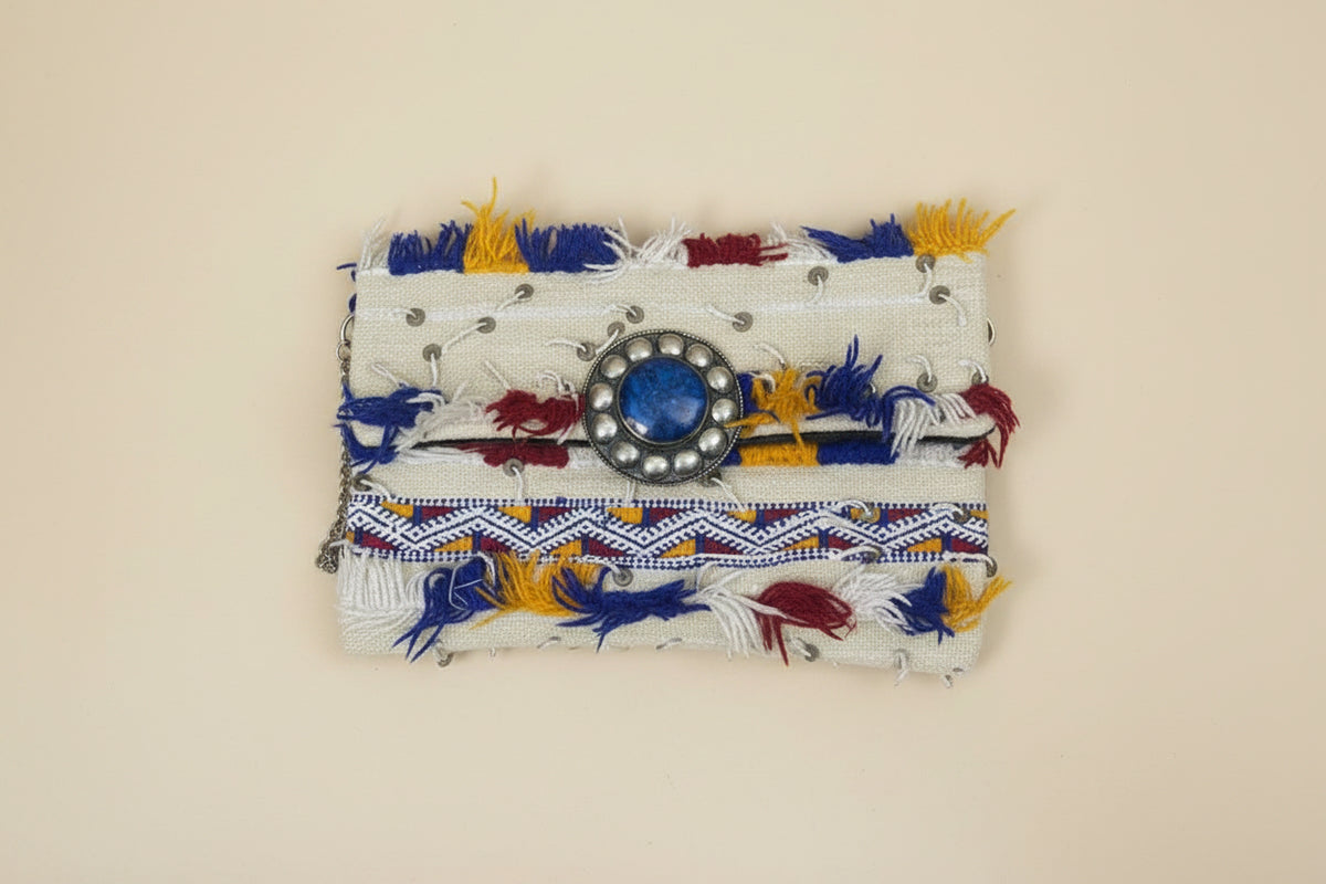 A small fabric clutch or wallet with a cream base, decorated with blue and yellow tassels, a blue stone in a silver-toned circular frame.