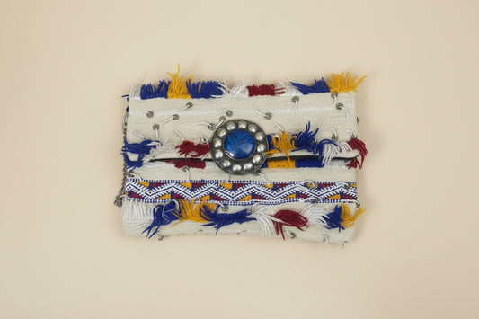 A small fabric clutch or wallet with a cream base, decorated with blue and yellow tassels, a blue stone in a silver-toned circular frame.